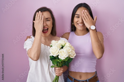 Hispanic mother and daughter holding bouquet of white flowers covering one eye with hand, confident smile on face and surprise emotion.