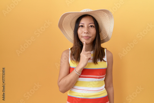Middle age chinese woman wearing summer hat over yellow background thinking concentrated about doubt with finger on chin and looking up wondering