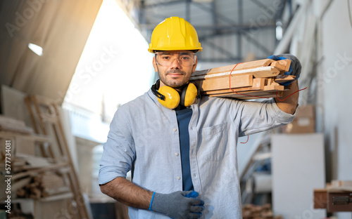 Male carpenter carrying wood planks on his shoulder. Portrait of young woodworker wearing safety helmet and gloves working on furniture renovation in carpenter's shop. Joiner, repairman at work.