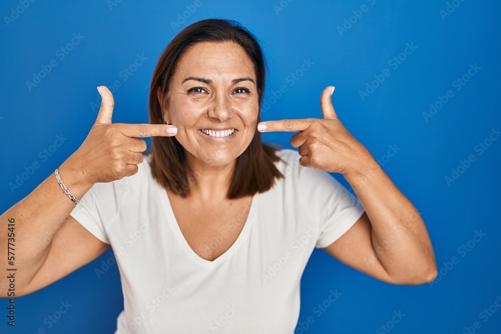 Hispanic mature woman standing over blue background smiling cheerful showing and pointing with fingers teeth and mouth. dental health concept. Hispanic mature woman standing over blue background smiling cheerful showing and pointing with fingers teeth and mouth. dental health concept.