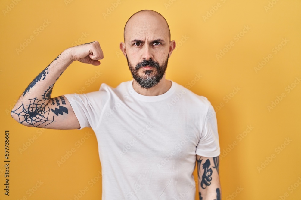 Young hispanic man with tattoos standing over yellow background strong person showing arm muscle, confident and proud of power Young hispanic man with tattoos standing over yellow background strong person showing arm muscle, confident and proud of power