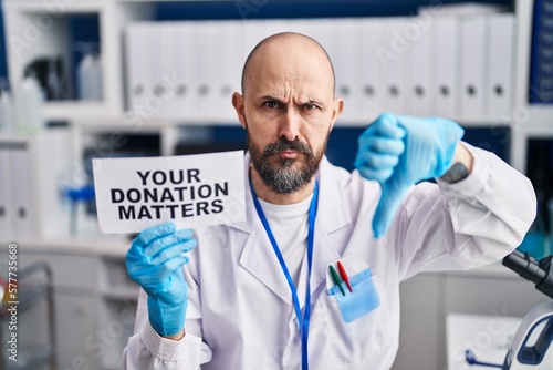 Young hispanic man working at scientist laboratory holding you donation matters banner with angry face, negative sign showing dislike with thumbs down, rejection concept