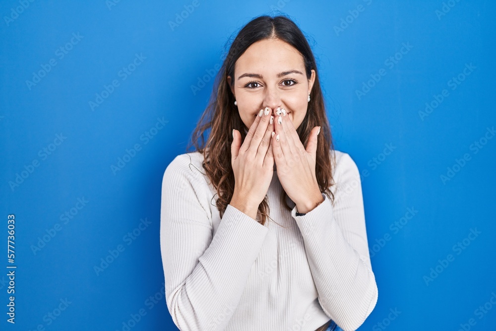 Young hispanic woman standing over blue background laughing and embarrassed giggle covering mouth with hands, gossip and scandal concept Young hispanic woman standing over blue background laughing and embarrassed giggle covering mouth with hands, gossip and scandal concept