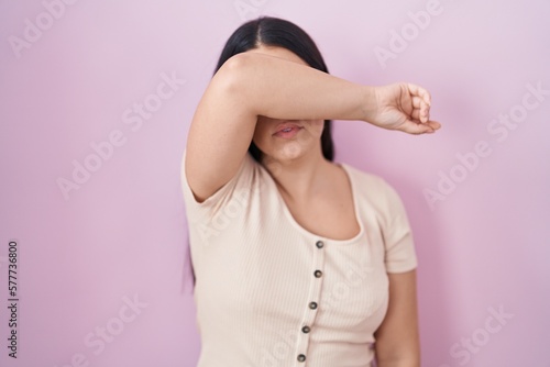 Young hispanic woman standing over pink background covering eyes with arm, looking serious and sad. sightless, hiding and rejection concept