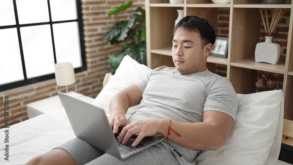 Young chinese man using laptop sitting on bed at bedroom