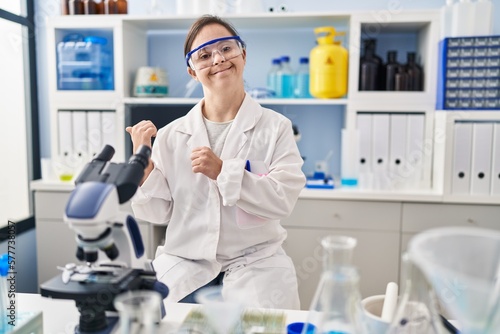 Hispanic girl with down syndrome working at scientist laboratory pointing to the back behind with hand and thumbs up, smiling confident