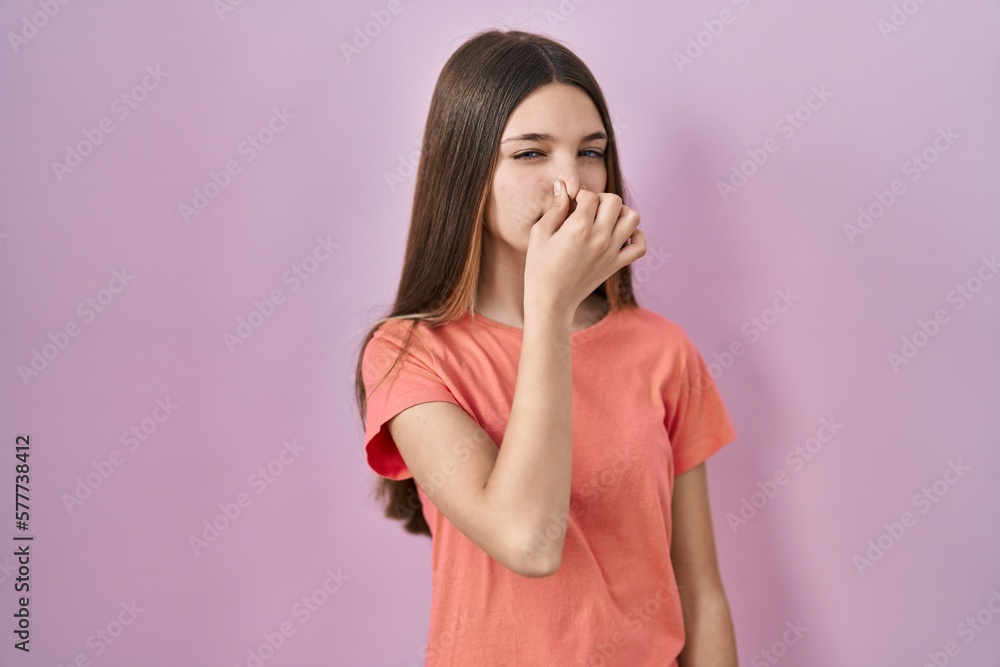 Teenager girl standing over pink background smelling something stinky and disgusting, intolerable smell, holding breath with fingers on nose. bad smell Teenager girl standing over pink background smelling something stinky and disgusting, intolerable smell, holding breath with fingers on nose. bad smell