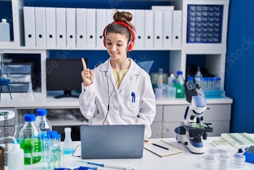Teenager girl working at scientist laboratory smiling with an idea or question pointing finger with happy face  number one