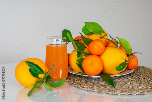 A glass of orange juice and fresh oranges and tangerines with leaves.