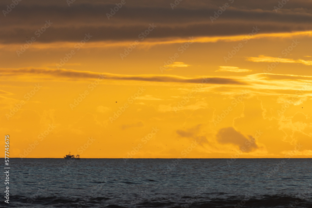A boat floats on the sea in the distance at a bright yellow sunset
