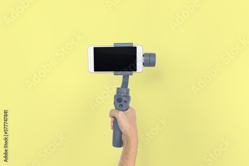 Hand holding gimbal or stabilizer with mobile phone on yellow pastel background. Vlog and travel concept. copy space. Video mobile.