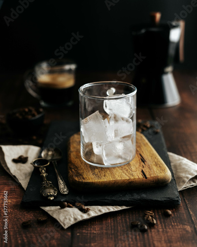 Taza de cristal transparente con hielo sobre una tabla de madera, jarra con café y fondo negro