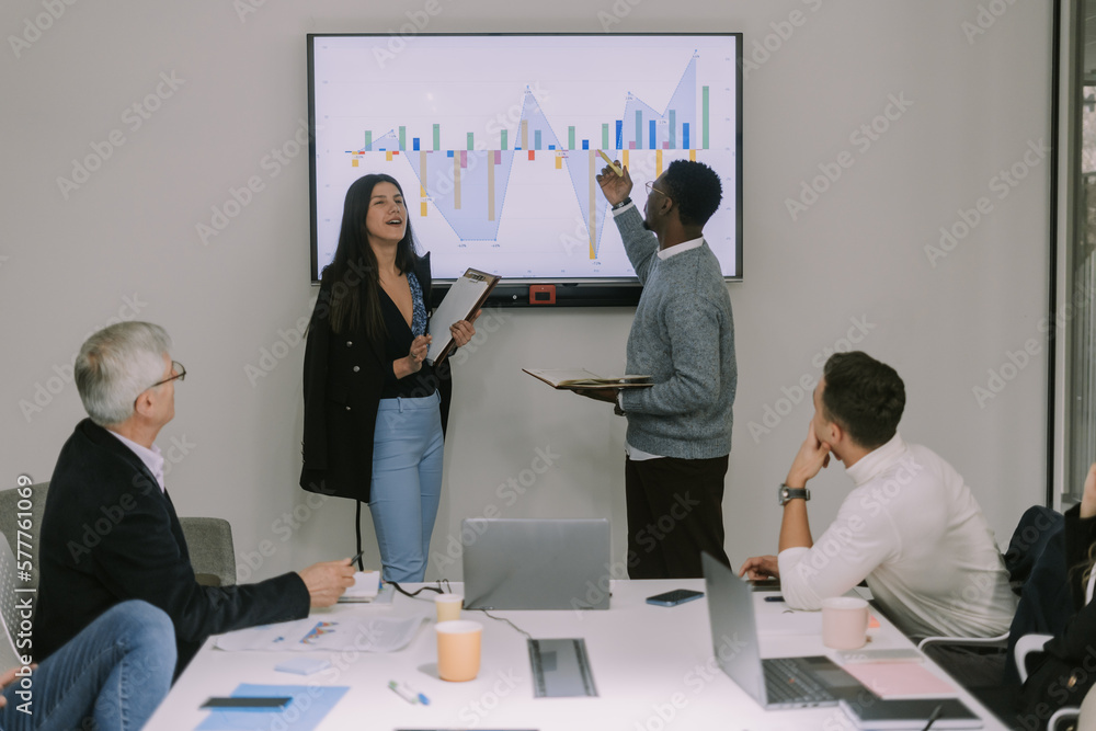 Black male pointing on the presentation monitor the same thing that his female partner is explaining to her colleagues Black male pointing on the presentation monitor the same thing that his female partner is explaining to her colleagues