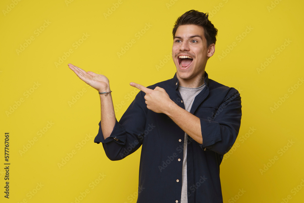 Young 20s brunette man exited smiling with left hand pointing to his opened right hand palm isolated on yellow background Young 20s brunette man exited smiling with left hand pointing to his opened right hand palm isolated on yellow background