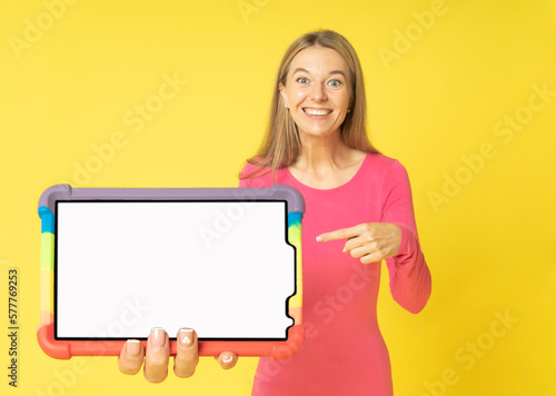Happy pretty woman showing or presenting tablet computer with white mock-up screen in hand and pointing isolated over yellow background. Beautiful woman holding big tablet pc in rainbow cover