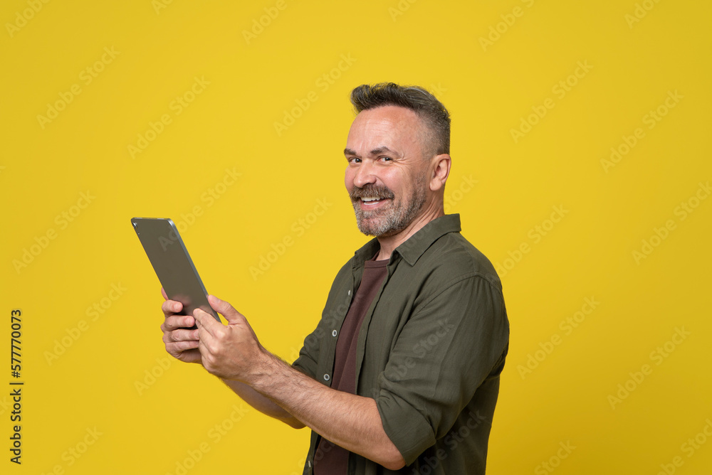 Mature man 50s dressed in green shirt holds digital tablet pc gadget in hands and happy smiled to camera after reading news or watching video isolated on yellow background Mature man 50s dressed in green shirt holds digital tablet pc gadget in hands and happy smiled to camera after reading news or watching video isolated on yellow background