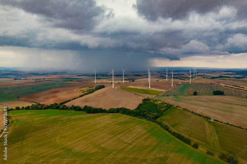 Drone flight at high altitude over a large field with many wind turbines spinning. The wind turbines cast a long shadow because the sun is setting. In the background, at storm sky.