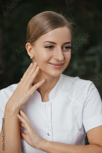 Smiling confident female doctor with lab coat on standing. Practician woman wearing white hospital uniform. Waist up portrait of young cosmetologist in white lab coat looking away with smile. 
