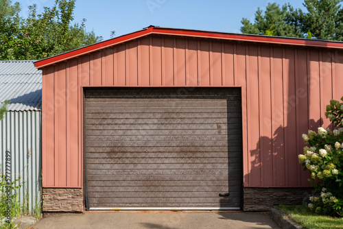 Modern red garage with driveway and roller door. Wide parking gate with shadows of trees and concrete driveway in front