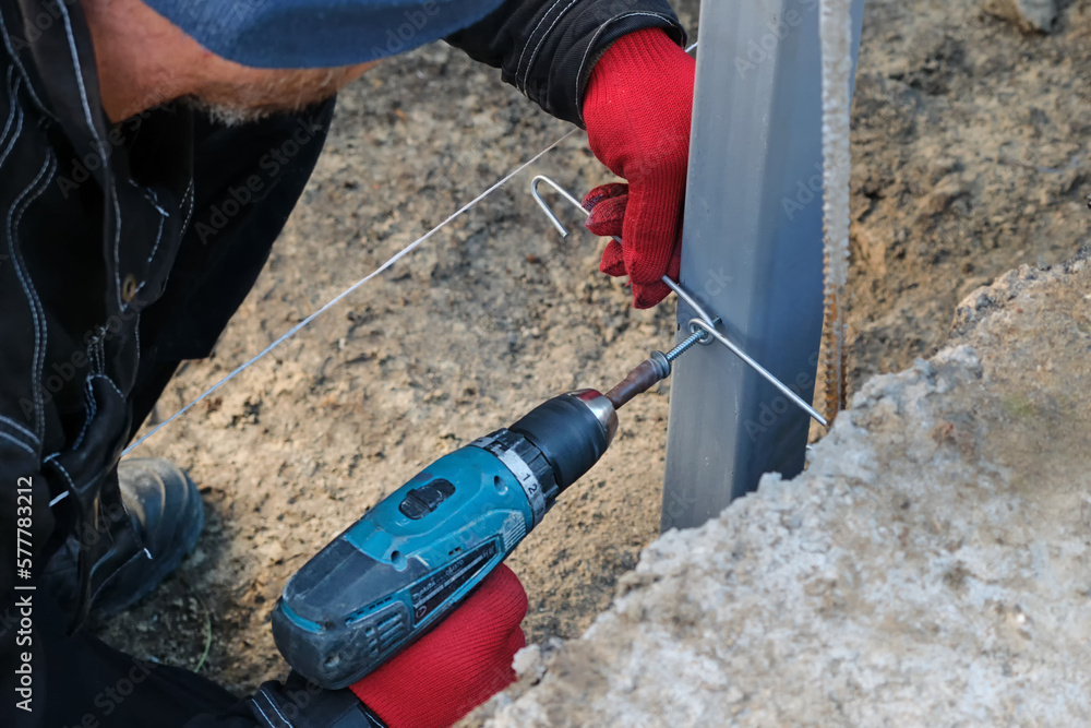 A worker in red gloves drilling a self-tapping screw into a steel post for mounting a fence.