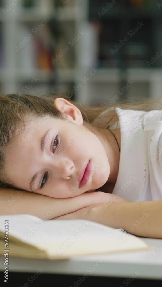 Vertical Screen: Sad girl lying on book and looking to side, pupil feeling tired or depressed, bookshelves on blurred background. Concept of education Vertical Screen: Sad girl lying on book and looking to side, pupil feeling tired or depressed, bookshelves on blurred background. Concept of education