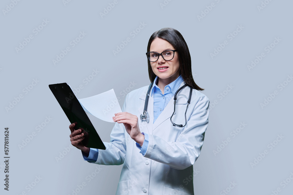 Young friendly woman doctor with clipboard on light gray studio background Young friendly woman doctor with clipboard on light gray studio background