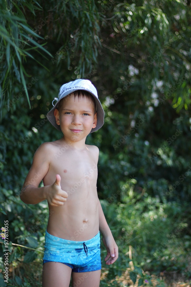 A white European schoolboy in swimming trunks and a hat shows a thumbs-up gesture against the backdrop of tropical foliage. Cool summer vacation. Copy space A white European schoolboy in swimming trunks and a hat shows a thumbs-up gesture against the backdrop of tropical foliage. Cool summer vacation. Copy space