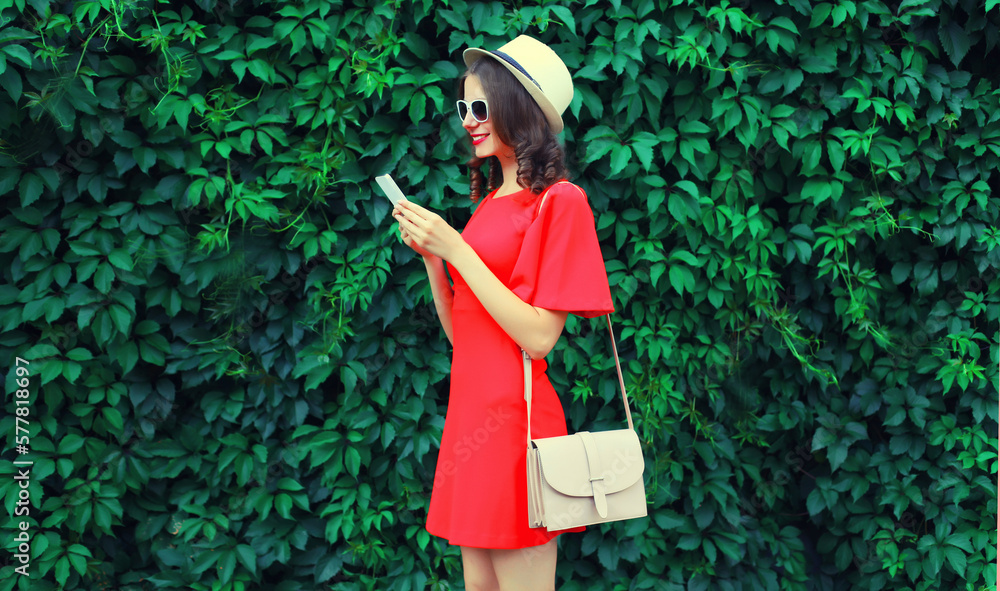 Portrait of happy smiling young woman with smartphone wearing summer straw hat, dress and handbag on green leaves wall background