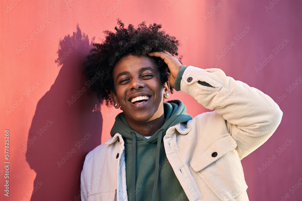 Young happy African American cool hipster guy face laughing on red city wall lit with sunlight. Smiling stylish cool rebel gen z teenager model standing outdoors. Headshot close up portrait shot. - obrazy, fototapety, plakaty 