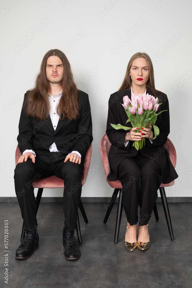 Obraz premium Elegant couple wearing black suits. A woman looking into the camera. A man holding a tulip and looking at it. Grey background. Place for your text.