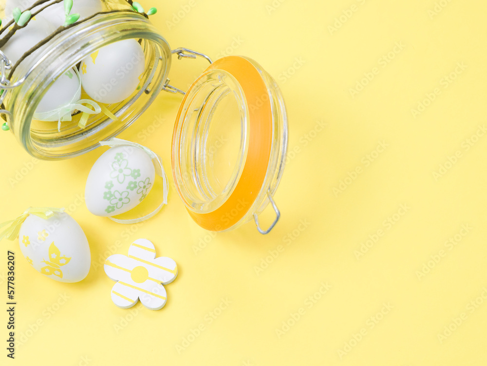 Decorative Easter eggs and a wooden flower pouring out of a lying glass jar on a yellow background