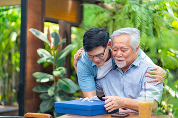 Asian man surprised elderly father with Birthday gift at outdoor cafe restaurant on summer holiday vacation. Family relationship, celebrating father's day and older people health care concept.