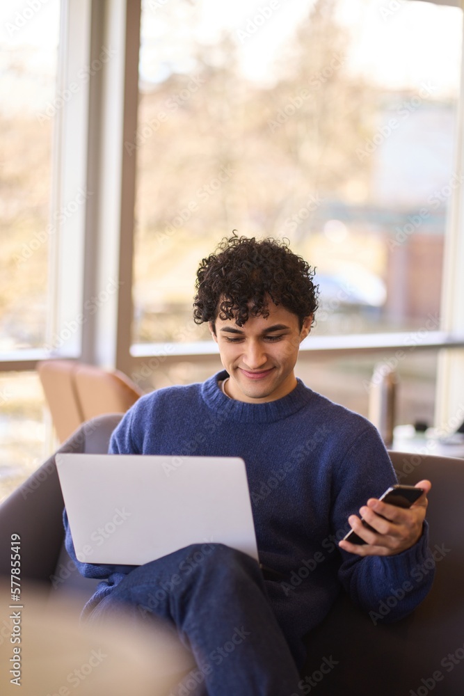 Hispanic young man, using mobile phone while working remotely on laptop in the office interior Hispanic young man, using mobile phone while working remotely on laptop in the office interior