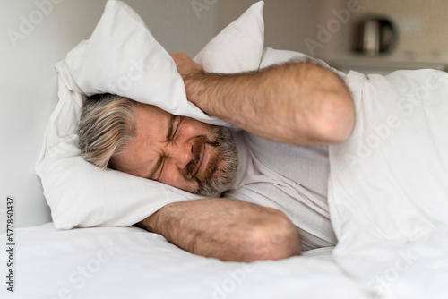 Irritated man lying in bed and covering ears with pillows