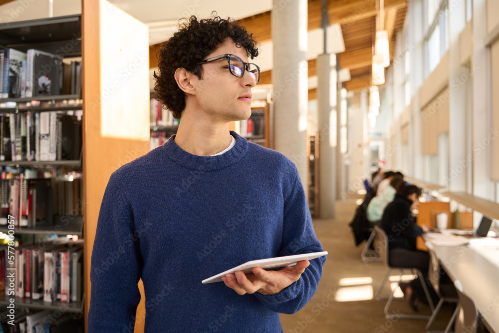 Fototapeta premium Smart Latin American young student in eyeglasses holding a tablet and looking aside in the library