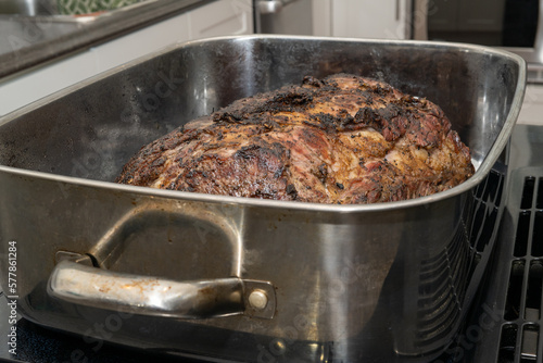 A large Angus beef prime rib roast cooked in a roaster pan with onions. The shiny metal pan is rectangular in shape with handles. The medium well done roast is covered in garlic.