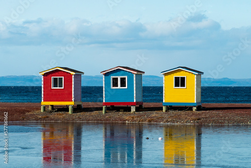 Three brightly colored storage sheds  red  blue  and yellow at the ocean s edge. There s a little pond in front of the buildings which is reflecting in the water. The sky is blue in the background.