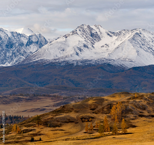 Russia, the Altai Mountains. View of the autumn mountains with a winding dirt road stretching into the distance against the background of the snow-covered North Chui mountain range.