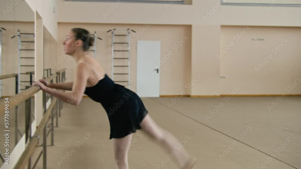 Female ballet dancer in training bodysuit stands near ballet barre, doing gymnastic leg movement, prepares for performance. Adult ballerina exercises in dance studio. School of classical ballet dance. Female ballet dancer in training bodysuit stands near ballet barre, doing gymnastic leg movement, prepares for performance. Adult ballerina exercises in dance studio. School of classical ballet dance.