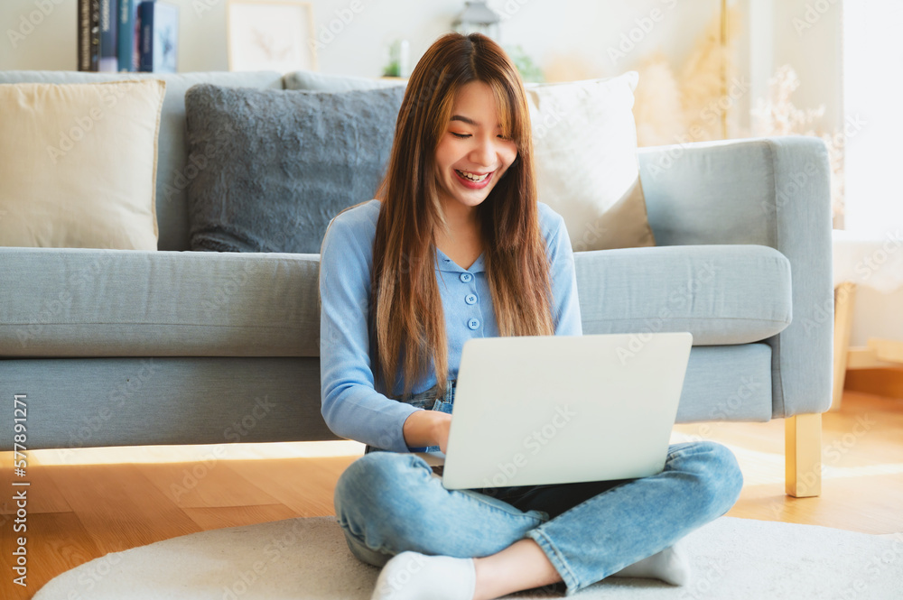 Young asian woman in good spirits working on laptop at home while sitting on the floor close to the couch