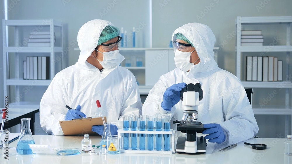Asian doctor scientist in biohazard protection clothing analyzing virus sample with microscope in laboratory, vaccine research. quarantine or virus outbreak concept Asian doctor scientist in biohazard protection clothing analyzing virus sample with microscope in laboratory, vaccine research. quarantine or virus outbreak concept