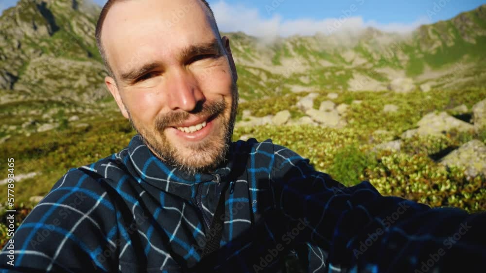 A travel blogger filming himself on a mountain hike. Face red from sunburn. A bearded Caucasian man with a short haircut smiles as he looks into the camera. Against a beautiful mountain backdrop A travel blogger filming himself on a mountain hike. Face red from sunburn. A bearded Caucasian man with a short haircut smiles as he looks into the camera. Against a beautiful mountain backdrop