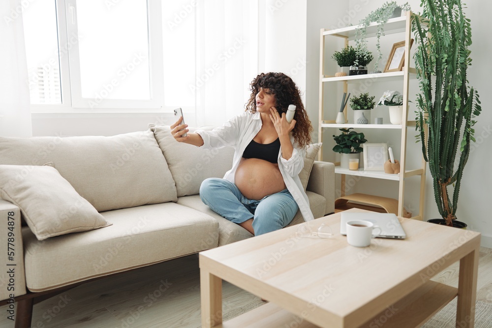 Pregnant woman smile blogger showing baby feeding bottle filming herself on phone, video chat sitting on sofa at home freelancer in last month of pregnancy lifestyle before childbirth Pregnant woman smile blogger showing baby feeding bottle filming herself on phone, video chat sitting on sofa at home freelancer in last month of pregnancy lifestyle before childbirth
