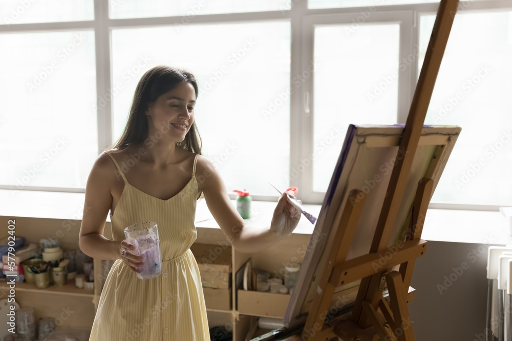 Happy inspired young female artist drawing artwork in creative studio, working at canvas on wooden easel, using water and paint brush, enjoying creativity, artistic hobby, smiling Happy inspired young female artist drawing artwork in creative studio, working at canvas on wooden easel, using water and paint brush, enjoying creativity, artistic hobby, smiling