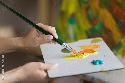 Palette of artist with acrylic paints close up. Art school student drawing picture in oil, combining yellow, orange colors on pallet, holding craft tools. Close up of hands