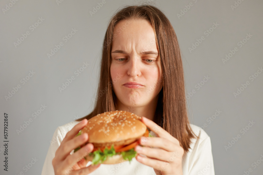 Portrait of dissatisfied sad woman wearing white T-shirt holding burger isolated on gray background, thinking eat or not, proper nutrition, healthy fast food, unhealthy choice.