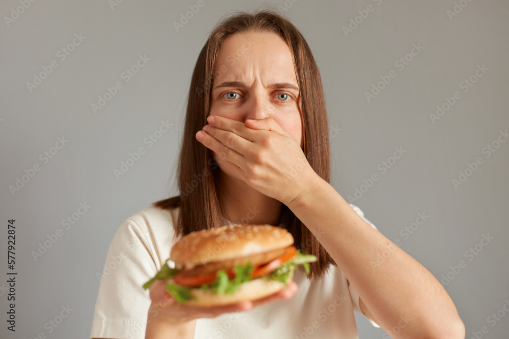 Indoor shot of ill woman feeling sick when see fast food, covering her mouth feels nausea, eating spoiled sandwich, looking at camera, feeling unwell.