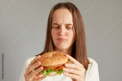 Portrait of dissatisfied sad woman wearing white T-shirt holding burger isolated on gray background, thinking eat or not, proper nutrition, healthy fast food, unhealthy choice.