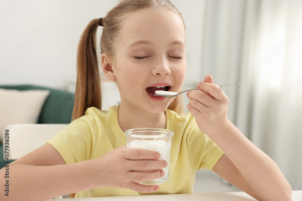 Cute little girl enjoying tasty yogurt at white table at home Cute little girl enjoying tasty yogurt at white table at home