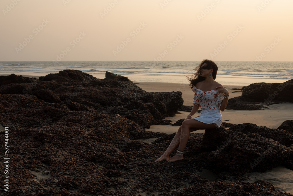Young woman with long hair fluttering in wind in short white skirt and blouse is sitting on rocks in sunset rays.